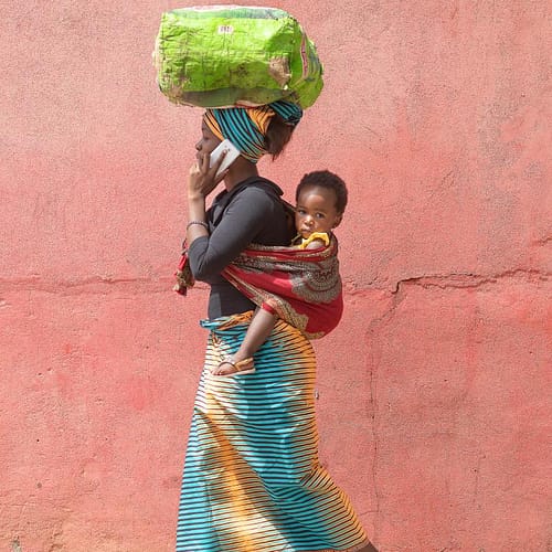 Moeder Florinda met baby Wanga op haar rug in een draagdoek loopt op straat in Mozambique met een rose muur op de achtergrond